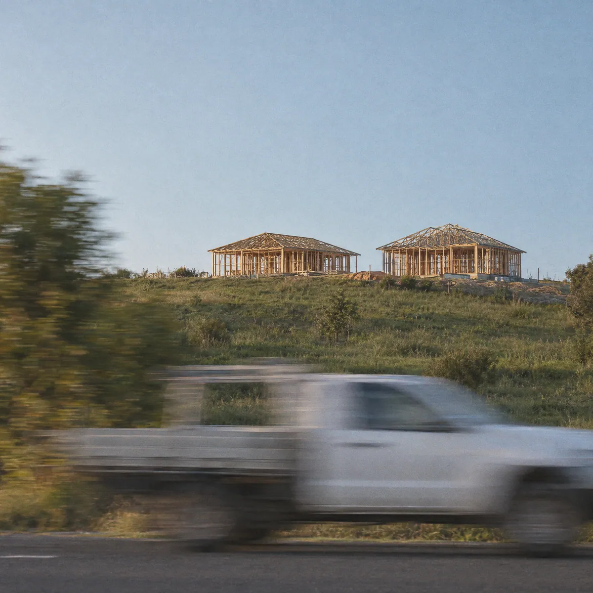 Two timber house frames on a grassy hill with a white ute streaked across the road below